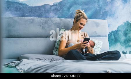 mamma bionda caucasica che allatta il bambino in camera da letto blu mentre guarda il suo telefono sorridente. Madre occupata. Responsabilità. Scatto orizzontale in interni. Foto di alta qualità Foto Stock