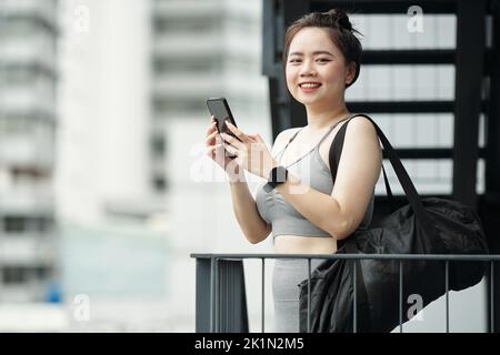 Ritratto di felice giovane donna in palestra vestiti in piedi sul balcone, testando pullman e sorridendo alla macchina fotografica Foto Stock