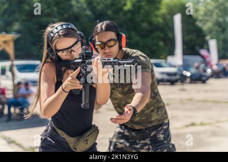 Un istruttore maschile muscolare aiuta una bella ragazza caucasica a maneggiare un fucile con un collimatore al campo di tiro. Foto di alta qualità Foto Stock