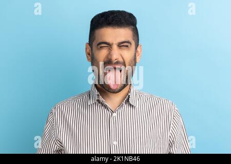 Foto di un giovane e stupido uomo d'affari pazzo, tenendo gli occhi chiusi e mostrando la lingua fuori, esprimendo emozioni positive, indossando una camicia a righe. Studio in interni isolato su sfondo blu. Foto Stock