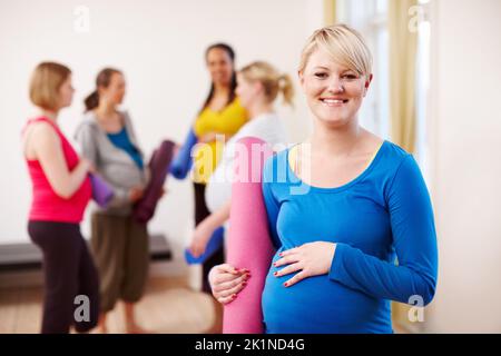 Pilates per la gravidanza. Una giovane donna bionda incinta in palestra con un materassino per esercizi e un gruppo di donne sullo sfondo. Foto Stock