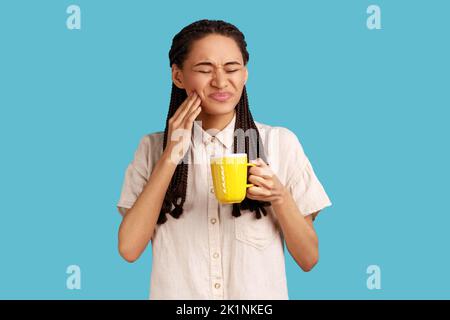 Ritratto di donna malsana con greggi neri che indossano camicia bianca. Tenere la tazza in mano, avendo dolore ai denti dopo aver bevuto bevande fredde o calde. Studio in interni isolato su sfondo blu. Foto Stock