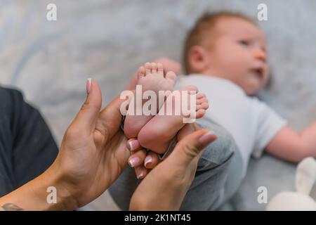 Una nuova madre amorevole che tiene dolcemente i suoi figli piccoli piedi. Un ragazzino dai capelli castani carini, adagiato sulla schiena su una coperta grigia con le braccia tese ai lati. Foto di alta qualità Foto Stock