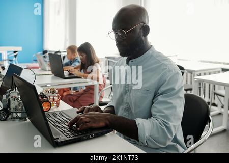 Serio giovane uomo nero in camicia casual seduto di fronte al notebook a lezione in classe contro due diligenti studentesse in rete dalla scrivania Foto Stock