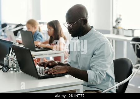 Giovane insegnante afro-americano serio di robotica seduto di fronte al computer portatile a lezione e in rete contro due scolaresche Foto Stock