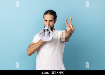 Ritratto di bell'uomo con barba che indossa una T-shirt bianca che tiene in mano il megafono e fa annuncio, mostrando il segno v alla fotocamera. Studio in interni isolato su sfondo blu. Foto Stock