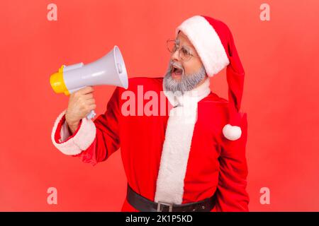 Ritratto di un uomo anziano pazzo con barba grigia che indossa il costume di babbo natale urlando forte, avendo un'espressione facciale arrabbiata, tenendo il megafono. Studio in interni isolato su sfondo rosso. Foto Stock