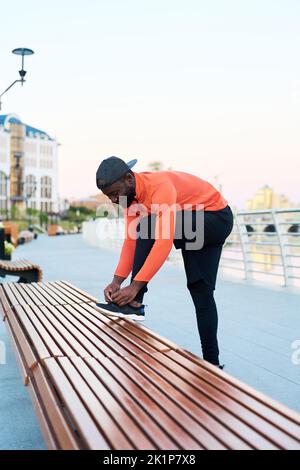 Vista laterale del giovane afro-americano in abbigliamento sportivo che lega le scarpe sulle sneaker mentre si piega sopra la gamba piegata nel ginocchio in strada Foto Stock