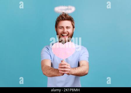 Ritratto dell'uomo angelico bearded con nimbus santo che tiene in mano il cuore di carta rosa sul bastone, dimostrante la cura di amore, sensibilità romantiche. Studio in interni isolato su sfondo blu. Foto Stock