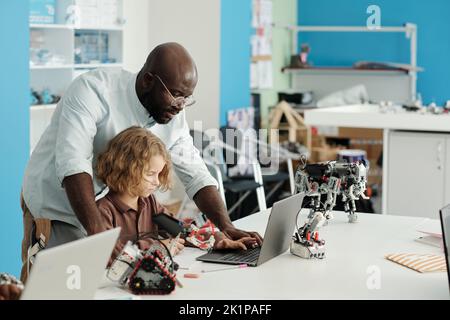 Giovane insegnante afroamericano di robotica che si piega su un simpatico scolaro seduto alla scrivania di fronte al computer portatile e che lavora su un nuovo progetto Foto Stock