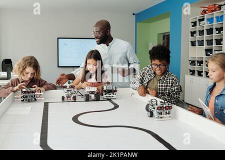 Giovane insegnante di robotica con un tablet che aiuta i ragazzi a controllare i loro robot durante il gioco dopo aver finito la creazione di nuovi oggetti Foto Stock