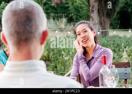 Giovane donna asiatica adulta sorridente e che parla con gli amici in una festa a cena seduta nel cortile. Concetto di stile di vita. Foto Stock