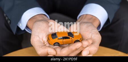 Mano d'uomo d'affari che tiene il giocattolo arancione dell'automobile sul tavolo. Finanziario, denaro, rifinanziamento e concetto di assicurazione auto Foto Stock