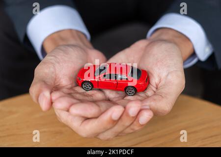 Mano d'uomo d'affari che tiene il giocattolo rosso dell'automobile sul tavolo. Finanziario, denaro, rifinanziamento e concetto di assicurazione auto Foto Stock