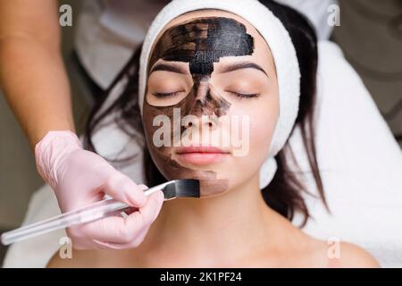 cosmetologo applicazione maschera nera sul viso bella donna indossare guanti neri, donna splendida in spa che hanno le procedure facciali Foto Stock