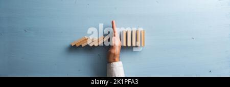 Mano di un uomo d'affari che smesso di crollare dominos per prevenire un disastro e una crisi. Vista dall'alto dello sfondo di legno blu. Foto Stock