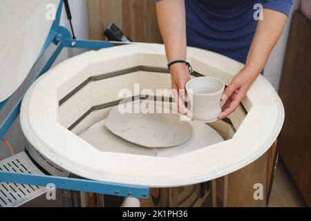 Primo piano delle mani mettere vaso nel forno, forno per tostare prodotti artigianali in argilla non cotta, ceramica non finita e arnese. Ceramica moderna Foto Stock