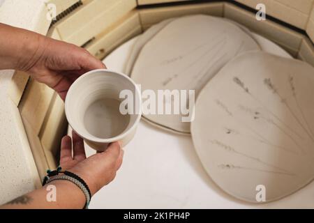 Primo piano delle mani mettere vaso nel forno, forno per tostare prodotti artigianali in argilla non cotta, ceramica non finita e arnese. Ceramica moderna Foto Stock