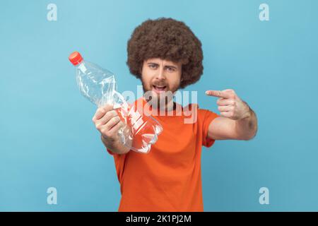 Ritratto di un bell'uomo con acconciatura afro che indossa una T-shirt arancione in piedi guardando la macchina fotografica, mostrando e puntando la bottiglia di plastica. Studio in interni isolato su sfondo blu. Foto Stock