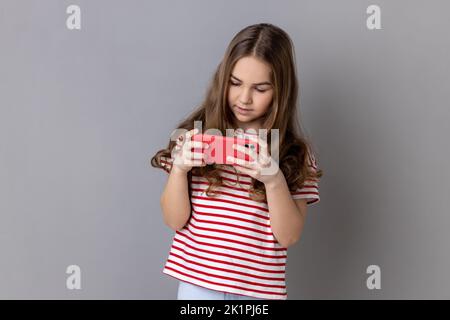 Animazione e tecnologia per bambini. Bambina in T-shirt a righe utilizzando il telefono cellulare, bambino che gioca a video gioco sul cellulare chattando in rete sociale. Studio in interni isolato su sfondo grigio Foto Stock