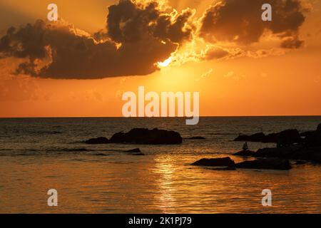 Tramonto e mare. Rocce o pietre sulla spiaggia. Cielo arancione. Sole nascosto dietro le nuvole. Silhouette. Orizzontale. Foto Stock