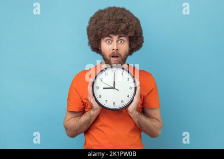 Ritratto dell'uomo scioccato con acconciatura afro in ritardo, tenendo in mano orologio da parete, scadenza, puntualità, guardando la fotocamera con bocca aperta. Studio in interni isolato su sfondo blu. Foto Stock