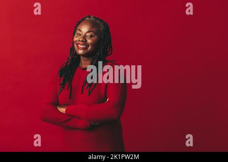Donna felice con i dreadlocks sorridente alla macchina fotografica mentre si trova su uno sfondo rosso con le braccia incrociate. Donna nera matura che abbraccia il suo natu Foto Stock