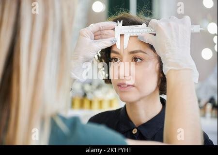 Estetista che utilizza lo strumento di misurazione per la procedura di correzione della fronte in salone di bellezza. Primo piano. Specialista di bellezza che posiziona il righello sulla fronte della donna mentre prende le misure per la correzione delle sopracciglia. Foto Stock