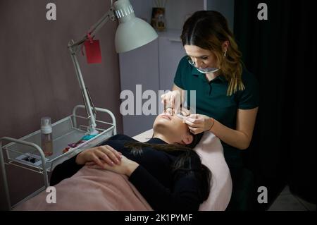 Specialista di bellezza femminile in maschera protettiva viso che pone occhio patch sul viso del cliente, mentre la donna sdraiata sul divano letto. Eyelash stilista preparare gli occhi di donna prima di applicare le estensioni di ciglia. Foto Stock