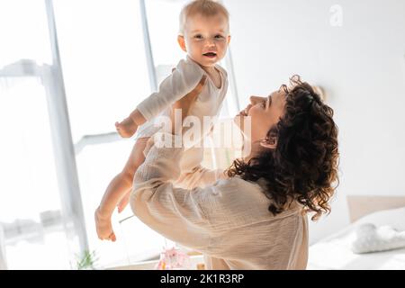 donna felice e riccia in loungewear tenendo in braccio nuda figlia in romper, immagine di scorta Foto Stock