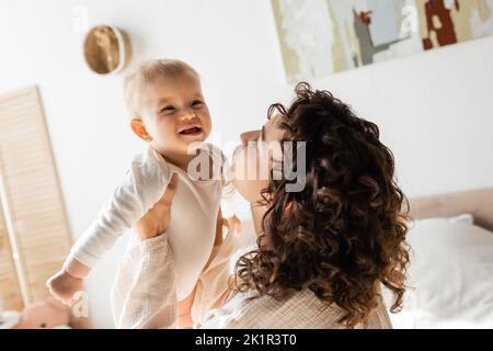 donna curly in loungewear tenendo in braccio bambino figlia in romper, immagine di scorta Foto Stock