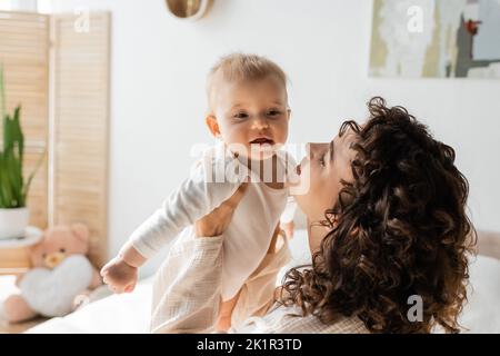 donna felice e riccia in loungewear tenendo in braccio bambino in romper, immagine di scorta Foto Stock