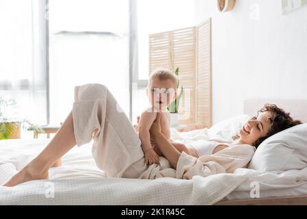 donna felice in loungewear sdraiato sul letto e tenendo bambino figlia, immagine di scorta Foto Stock