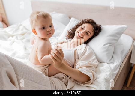 donna felice in loungewear sdraiato sul letto e tenendo la mano della figlia del bambino, immagine di scorta Foto Stock
