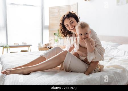 donna allegra in loungewear tenendo in braccio bambino e seduta sul letto, immagine stock Foto Stock