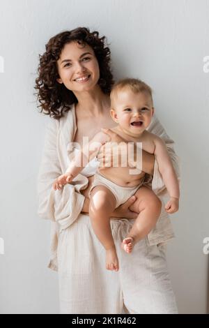 Mamma bruna positiva in pigiama tenendo la bambina a casa, immagine stock Foto Stock