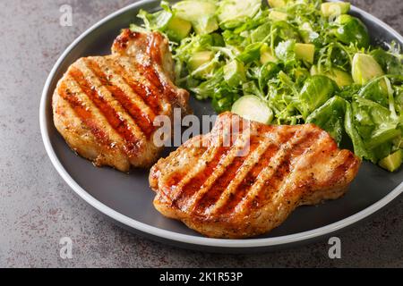 Bistecca di maiale su un osso e insalata verde con germogli di bruxelles, avocado e erbe closeup nel piatto sul tavolo. Orizzontale Foto Stock