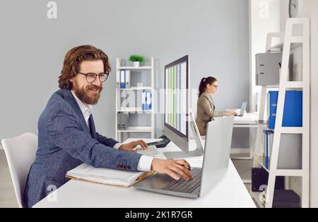 Allegro contabile maschile in ufficio che lavora con database utilizzando il computer portatile e libro di contabilità. Foto Stock