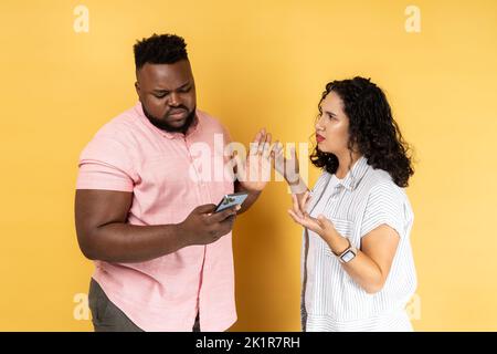 Giovane coppia in abbigliamento casual in piedi insieme, l'uomo che utilizza il telefono cellulare e ignorare sua moglie, donna cercando di attirare la sua attenzione e urlare. Studio al coperto isolato su sfondo giallo. Foto Stock