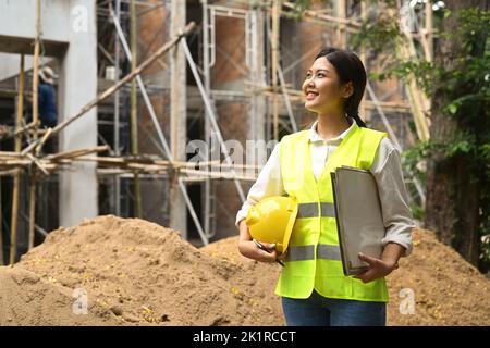 Donna architetto premuroso in giacche riflettenti che tiene hardhat e blueprints in piedi davanti al sito di costruzione di edifici commerciali Foto Stock