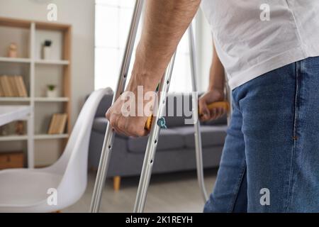 L'uomo adulto con infortunio cammina intorno all'appartamento a casa, appoggiandosi alle moderne stampelle metalliche. Foto Stock