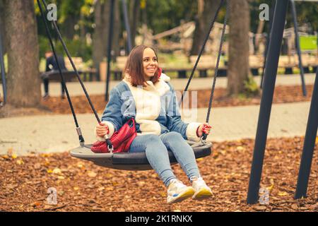 Una bella e allegra donna in abiti caldi oscilla su un altalena nel parco. Ragazza che dondola nel parco cittadino autunnale. Foto Stock