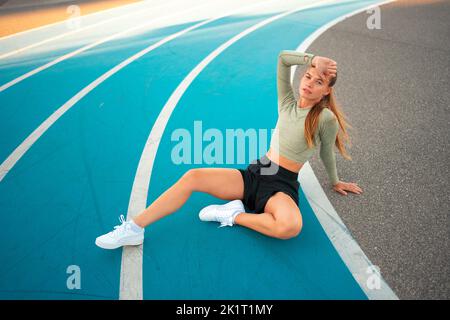 Stanco arrabbiato atleta femminile seduta sulla pista di corsa riposo dopo esercizi di jogging. Risultato sportivo deluso. Sport dilettante sprinter esaurito dopo allenamenti impegnativi seduti in pista Foto Stock