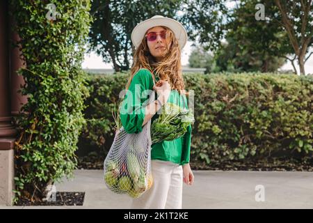 Ritratto di una giovane donna che tiene una borsa a rete con veggie sulla spalla mentre cammina per strada Foto Stock