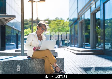 Felice e attraente donna d'affari di mezza età che lavora sorridendo con un computer portatile di fronte a un edificio moderno Foto Stock