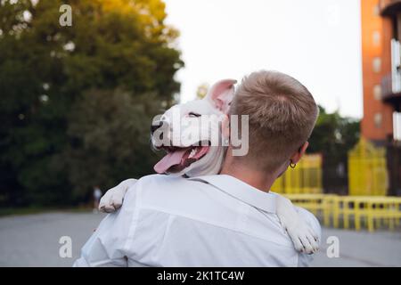 Il proprietario del cane abbraccia il suo giovane cane all'aperto. Uomo che interagisce con un cucciolo di terrier bianco dello staffordshire, felicità, gioia ed emozioni positive con gli animali domestici Foto Stock