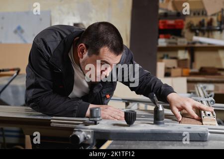 Vista laterale ritratto di un lavoratore maschio che utilizza una macchina per il taglio del legno in officina e che misura con attenzione Foto Stock