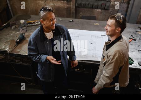 Ritratto ad angolo alto di due sorridenti operai di fabbrica che discutono il progetto mentre sono in officina Foto Stock