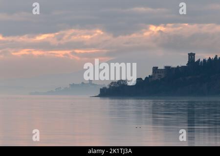 Vista tranquilla sul lago Trasimeno al tramonto, con isole ed edifici che si silano attraverso una leggera nebbia Foto Stock