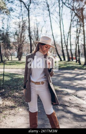 Donna sottile e graziosa che indossa blazer, cappello e stivali eleganti tenendo la borsa nel parco. Abito femminile autunnale alla moda. Look autunnale alla moda Foto Stock